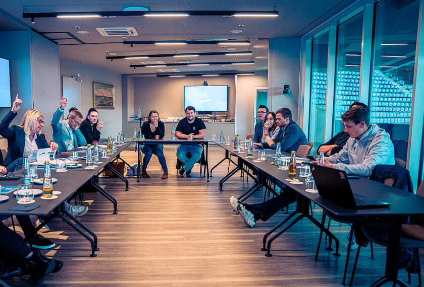 Eine Gruppe von Menschen sitzt in einem modernen Besprechungsraum an Tischen, die zu einer langen Reihe verbunden sind. Einige Teilnehmer heben die Hände, während andere aufmerksam zuhören oder sich Notizen machen. Auf den Tischen stehen Wasserflaschen und Gläser. Im Hintergrund ist eine Präsentation auf einem Bildschirm zu sehen, und die Wände sind mit großen Fenstern und einem Regal mit Flaschen gestaltet.