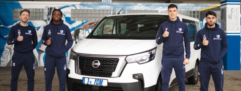 Vier Fußballspieler in blauen Trainingsanzügen stehen vor einem weißen Nissan-Fahrzeug. Sie zeigen alle mit Daumen nach oben und lächeln in die Kamera. Im Hintergrund ist eine Wand mit Graffiti und Bildern von Fußballspielern zu sehen.