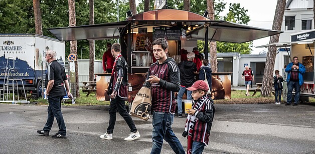 Ein Mann und ein Kind gehen auf einem Gehweg vorbei, im Hintergrund ist ein Imbissstand mit mehreren Personen zu sehen.