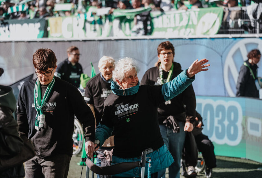 Eine ältere Frau im Rollstuhl wird von einem jüngeren Mann begleitet, während sie auf dem Fußballfeld einlaufen. Beide tragen schwarze T-Shirts mit der Aufschrift 'Fairer Miteinander' und grüne Schals. Im Hintergrund sind Fans mit grünen Schals und Transparenten zu sehen, die die Szene beobachten. Die Atmosphäre wirkt inklusiv und vielfältig.