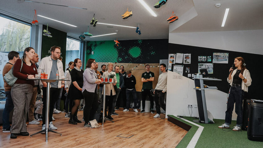 Eine Gruppe von Menschen steht in einem modernen Raum mit grünem Boden und einer schwarzen Tafel im Hintergrund. Einige Personen halten Getränke in der Hand und unterhalten sich an einem Stehtisch. Eine Person steht an einem Podium und scheint eine Präsentation oder einen Vortrag zu halten. Der Raum ist mit verschiedenen Postern und Bildern dekoriert, und es gibt eine Projektionsfläche an der Wand.