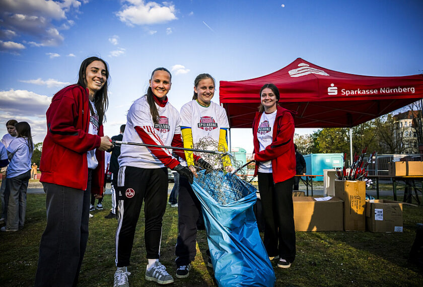 Vier Frauen in Sportkleidung stehen auf einem Rasenfeld und halten gemeinsam eine blaue Müllsammeltasche. Sie lächeln in die Kamera. Im Hintergrund ist ein roter Pavillon mit dem Logo der Sparkasse Nürnberg zu sehen.