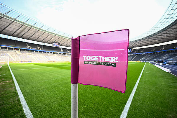 Eine pinkfarbene Fahne mit der Aufschrift 'TOGETHER! STOP HATE, BE A TEAM.' steht auf einem Fußballfeld in einem Stadion. Im Hintergrund sind die leeren Sitzplätze und das Dach des Stadions zu sehen.