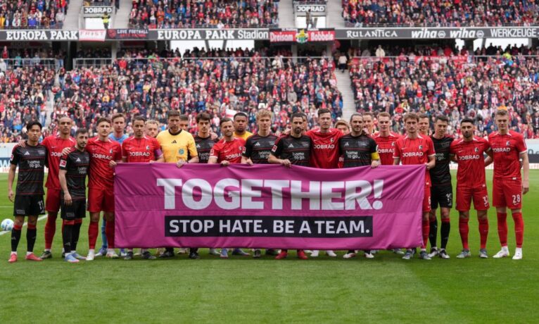 Zwei Fußballmannschaften stehen auf dem Rasen des Stadions und halten gemeinsam ein großes pinkes Transparent mit der Aufschrift "TOGETHER! STOP HATE. BE A TEAM." in die Höhe. Im Hintergrund sind zahlreiche Zuschauer auf den Rängen zu sehen.
