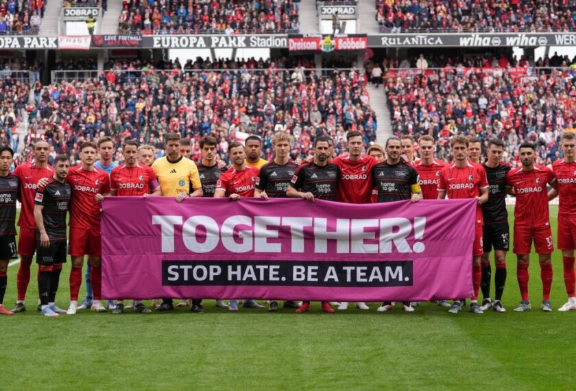 Zwei Fußballmannschaften stehen auf dem Rasen des Stadions und halten gemeinsam ein großes pinkes Transparent mit der Aufschrift "TOGETHER! STOP HATE. BE A TEAM." in die Höhe. Im Hintergrund sind zahlreiche Zuschauer auf den Rängen zu sehen.