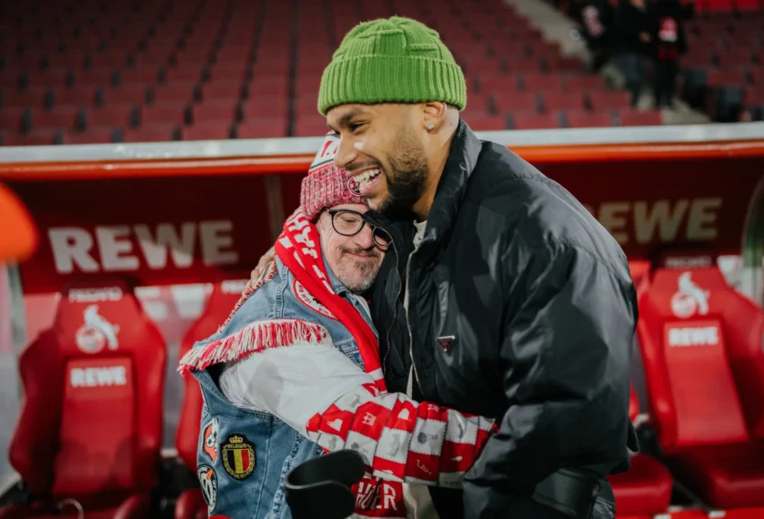 Zwei Personen umarmen sich herzlich in einem Fußballstadion. Eine Person trägt eine grüne Mütze und eine schwarze Jacke, während die andere Person eine rote und weiße Schärpe sowie eine blaue Jacke mit Aufnähern trägt. Im Hintergrund sind rote Sitzplätze und Werbebanner mit der Aufschrift "REWE" zu sehen.