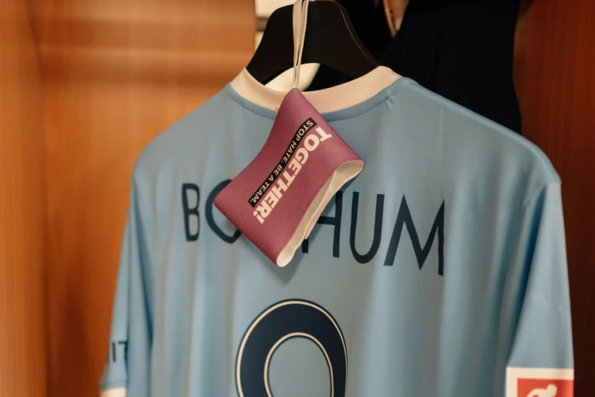 Ein hellblaues Trikot hängt an einem Bügel. Auf dem Trikot steht der Schriftzug "BOCHUM" und darunter ein Logo. An dem Bügel ist ein pinkfarbenes Schild befestigt, auf dem "TOGETHER: STOP HATE BE A TEAM" steht.