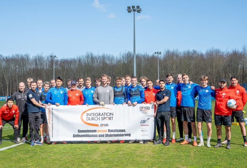 Eine Gruppe von Menschen steht auf einem Fußballfeld und hält ein Banner mit der Aufschrift "Integration durch Sport" und "Gemeinsam gegen Rassismus, Antisemitismus und Diskriminierung". Die Personen tragen Sportkleidung in verschiedenen Farben, darunter blaue, rote und schwarze Outfits. Im Hintergrund sind Bäume und ein Zaun mit Werbung zu sehen.