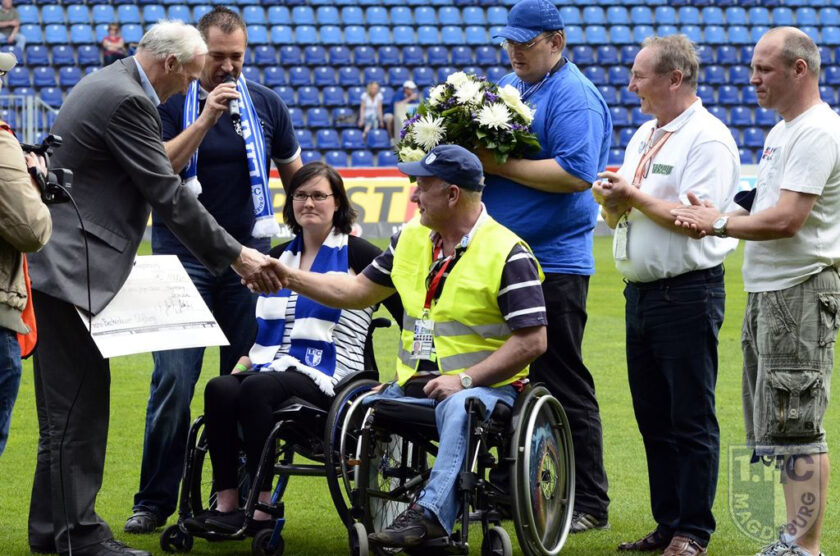 Ein Mann im Rollstuhl schüttelt einem anderen Mann die Hand, während er einen Blumenstrauß hält. Beide sind von einer Gruppe von Menschen umgeben, die auf einem Fußballfeld stehen. Der Mann im Rollstuhl trägt eine gelbe Weste und eine Mütze, während der andere Mann einen Anzug trägt. Die Gruppe scheint an einer offiziellen Veranstaltung teilzunehmen, möglicherweise im Zusammenhang mit einem Fußballspiel oder einer Fußballveranstaltung.