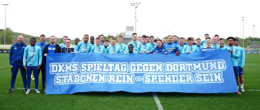 Eine Gruppe von Fußballspielern und Trainern posiert auf einem Rasenfeld für ein Gruppenfoto. Sie tragen blaue Trainingskleidung und halten ein großes blaues Banner mit der Aufschrift "DKMS Spieltag gegen Dortmund - Stäbchen rein - Spender sein". Die Spieler und Trainer lächeln und stehen dicht beieinander, was eine gemeinsame Aktion oder Kampagne unterstützt.