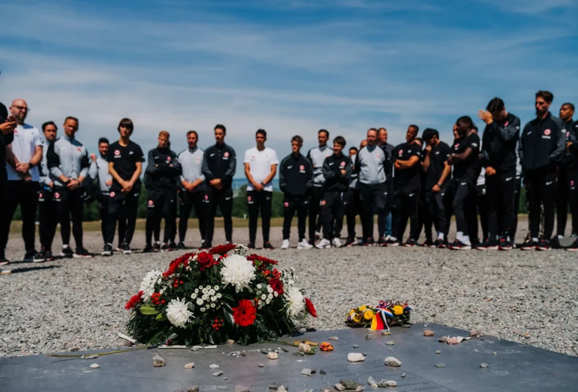 Eine Gruppe von Menschen steht in einer Reihe und hält eine Gedenkzeremonie ab. Vor ihnen liegt ein Kranz mit roten und weißen Blumen auf einem grauen Stein, der mit kleinen Steinen bedeckt ist. Im Hintergrund ist eine offene Landschaft mit Himmel zu sehen.