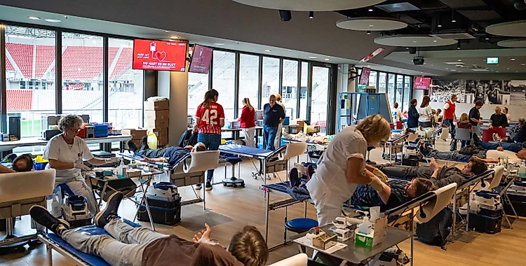 Eine Blutspendeaktion in einem Stadion. Menschen liegen auf Liegen und spenden Blut, während medizinisches Personal sie betreut. Im Hintergrund sind Tribünen und eine große Fensterfront zu sehen.