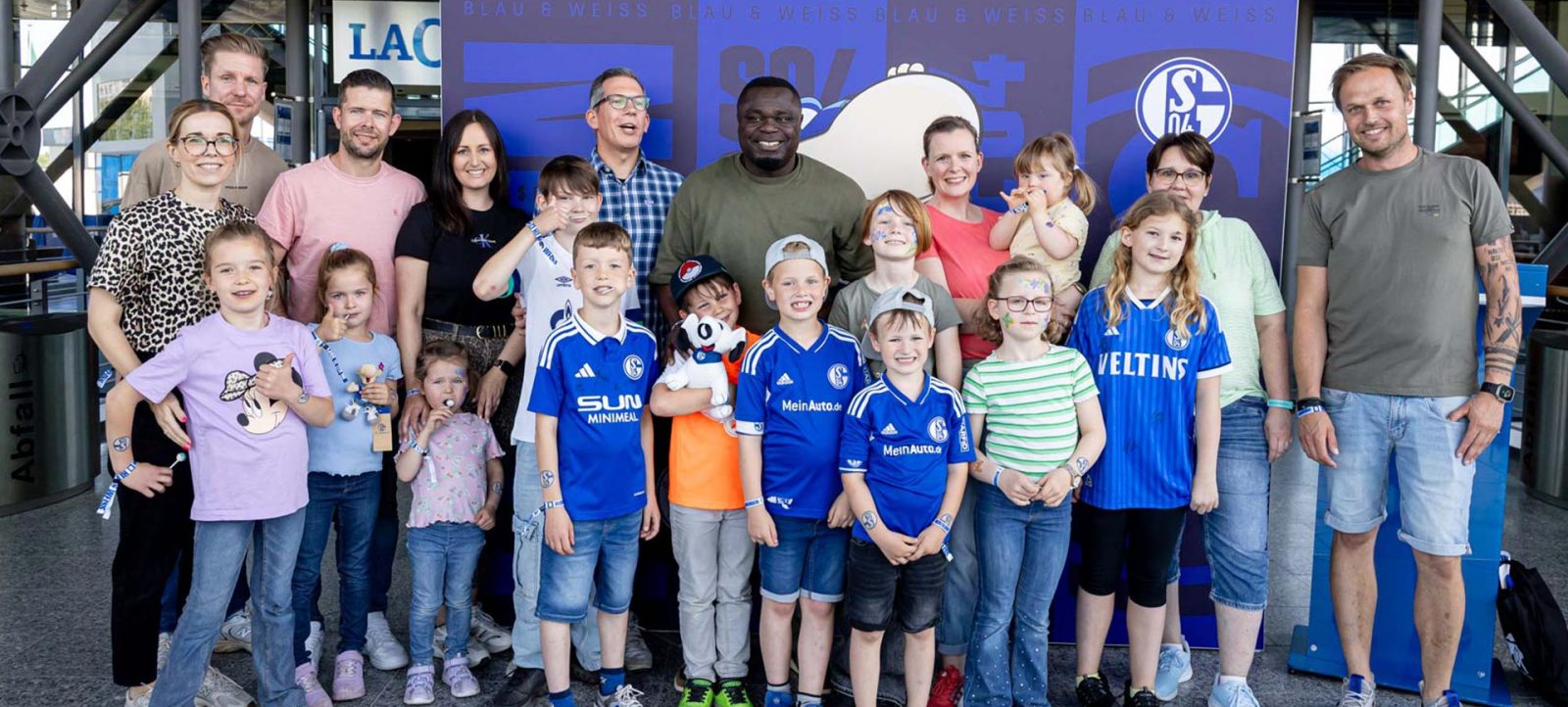Eine große Gruppe von Menschen, darunter Kinder und Erwachsene, steht vor einem blauen Hintergrund mit dem Logo des FC Schalke 04. Die Menschen tragen verschiedene T-Shirts, einige mit dem Logo des FC Schalke 04. Sie lächeln und posieren für ein Gruppenfoto.