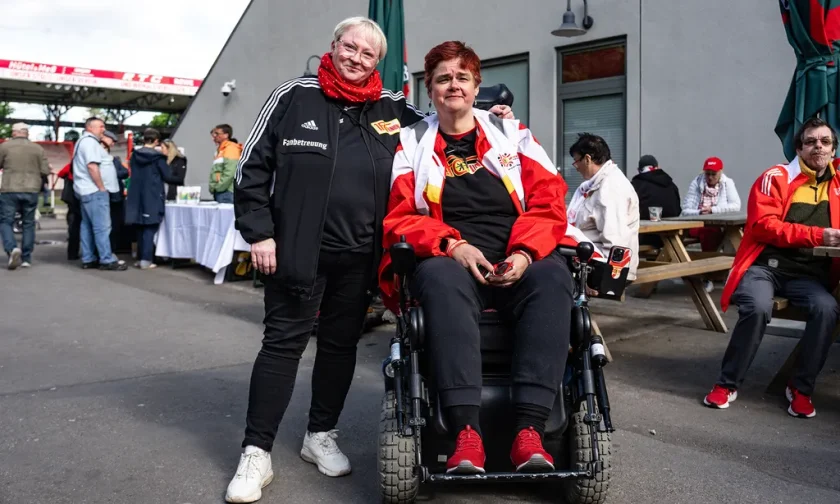 Zwei Personen stehen vor einem Gebäude, eine davon in einem Rollstuhl. Die Person im Rollstuhl trägt ein T-Shirt mit einem Vereinslogo und eine rote Jacke. Die andere Person trägt eine schwarze Adidas-Jacke mit der Aufschrift "Fansbetreuung" und einen roten Schal. Im Hintergrund sind weitere Menschen zu sehen, die sich unterhalten oder an Tischen sitzen.