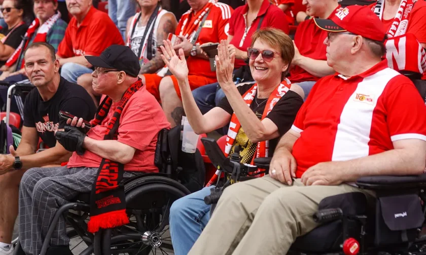 Eine Gruppe von Menschen, darunter einige in Rollstühlen, sitzen in einem Stadion und feiern begeistert. Sie tragen rote und schwarze Kleidung und Accessoires, die mit dem Logo "Borussia Dortmund" verziert sind. Die Atmosphäre ist fröhlich und energiegeladen, während sie gemeinsam den Moment genießen.