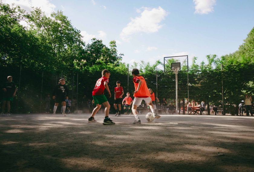 Zwei Kinder spielen auf einem staubigen Bolzplatz Fußball. Die Kinder tragen rote Trikots und orange Trainingswesten. Im Hintergrund sind weitere Kinder und Erwachsene zu sehen, die dem Spiel zuschauen. Der Bolzplatz ist von einem Zaun umgeben und befindet sich in einem Park mit vielen Bäumen.