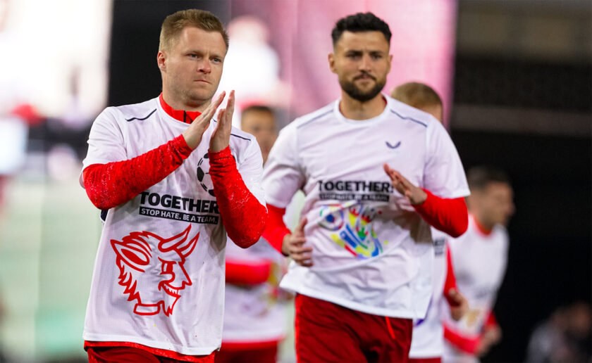 Zwei Fußballspieler des 1. FC Kaiserslautern klatschen sich auf dem Spielfeld ab. Sie tragen weiße Trikots mit roten Ärmeln und einem Aufdruck, der für Zusammenhalt und gegen Hass wirbt. Im Hintergrund ist ein unscharfes Stadion zu sehen.