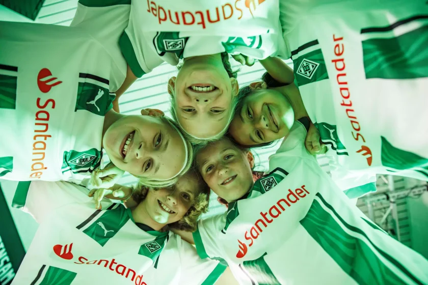 Vier Kinder lächeln in die Kamera, während sie sich in der Mitte eines Kreises aus grün-weißen Fußballtrikots mit dem Logo von 1. FC Köln und dem Sponsor "Santander" befinden.