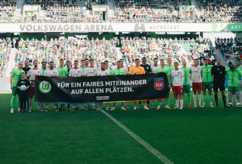 Zwei Fußballmannschaften stehen vor einem Spiel in der Volkswagen Arena. Sie halten ein Banner mit der Aufschrift 'Für ein faires Miteinander auf allen Plätzen.' im Stadion. Die Spieler tragen ihre Teamtrikots und posieren für ein Gruppenfoto. Im Hintergrund sind zahlreiche Zuschauer auf den Tribünen zu sehen.