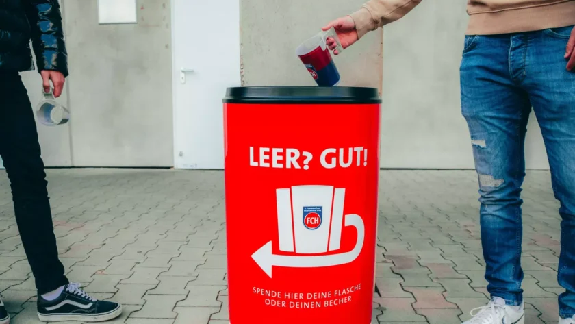 Zwei Personen werfen leere Flaschen in einen roten Müllcontainer mit der Aufschrift 'LEER? GUT!' und 'Spende hier deine Flasche oder deinen Becher'. Der Container hat ein Logo mit den Buchstaben 'FCH' und einen Pfeil, der nach unten zeigt.
