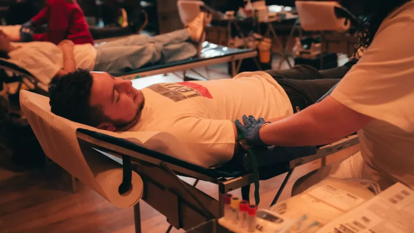 Ein Mann liegt auf einer Liege und spendet Blut. Sein linker Arm ist mit einem Schlauch verbunden, der das Blut in eine Sammelvorrichtung leitet. Im Hintergrund sind weitere Personen zu sehen, die ebenfalls Blut spenden. Auf dem Tisch vor der Liege befinden sich verschiedene Utensilien und Behälter für die Blutspende.