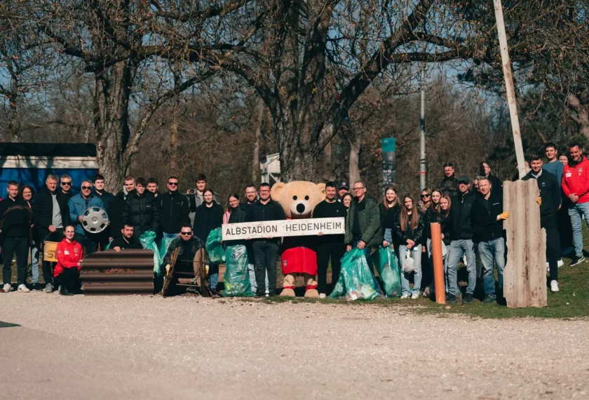 Eine Gruppe von Menschen, darunter auch eine Maske des Vereins-Maskottchens, steht vor dem Albstadion in Heidenheim. Sie halten Müllsäcke in den Händen und scheinen an einer Umweltaktion teilzunehmen. Im Hintergrund sind Bäume und ein Teil eines Fahrzeugs zu sehen.