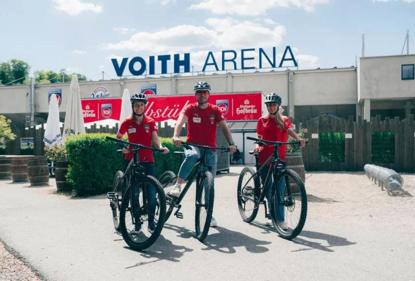 Drei Personen mit Fahrrädern vor der Voith Arena. Die Personen tragen rote T-Shirts und Helme und lächeln in die Kamera. Im Hintergrund ist die Fassade der Arena mit Werbebannern zu sehen.