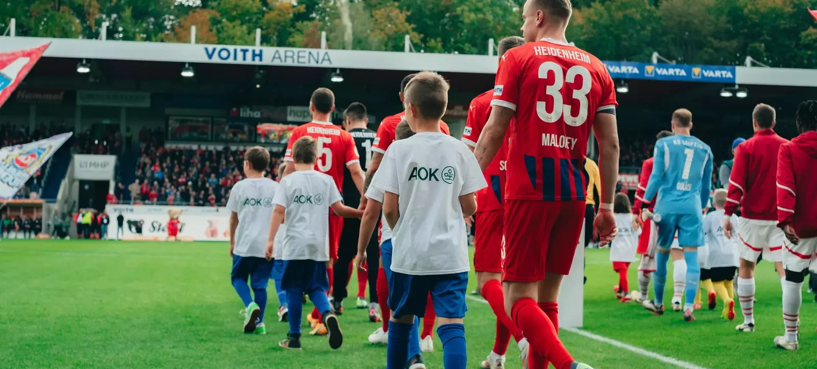 Spieler und Kinder laufen auf dem Fußballfeld der Voith Arena. Die Spieler tragen rote Trikots, während die Kinder weiße T-Shirts mit dem Logo "AOK" auf dem Rücken tragen. Im Hintergrund sind die Tribünen mit Zuschauern zu sehen.