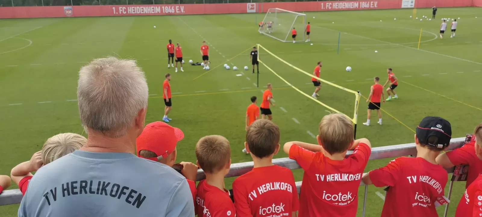 Eine Gruppe von Kindern und einem Erwachsenen beobachtet ein Fußballtraining auf dem Platz des 1. FC Heidenheim 1846 e.V. Die Zuschauer tragen T-Shirts mit dem Aufdruck "TV Herlikofen" und "icotek". Die Spieler auf dem Feld sind in roten Trikots und schwarzen Hosen gekleidet und führen verschiedene Übungen aus. Im Hintergrund ist die Werbeband mit dem Vereinsnamen zu sehen.