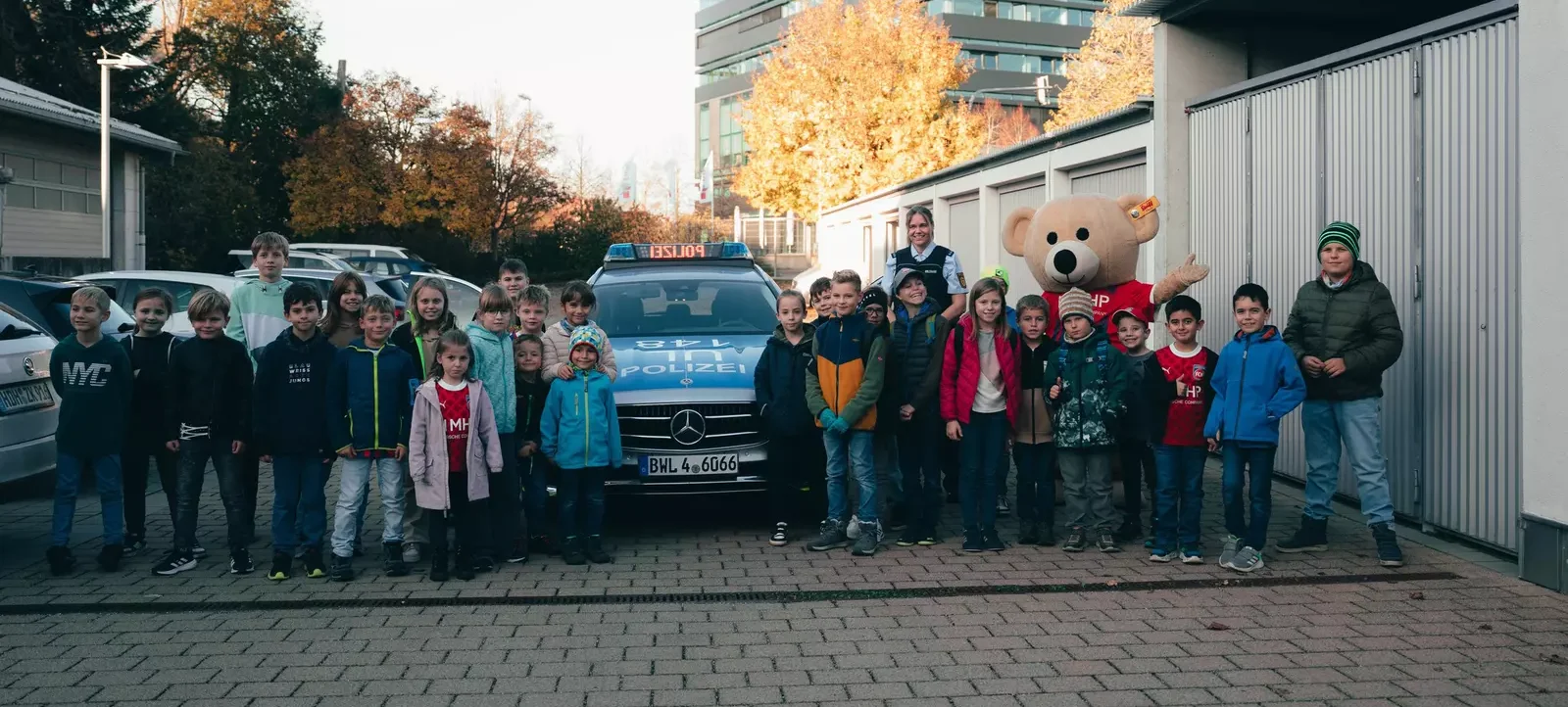 Eine Gruppe von Kindern und Erwachsenen steht vor einem Polizeiauto und posiert für ein Gruppenfoto. Ein großer Teddybär in einem roten T-Shirt mit der Aufschrift "HP" steht in der Mitte der Gruppe. Die Kinder und Erwachsenen tragen verschiedene Jacken und Kleidung und scheinen sich vor einem Gebäude mit grauen Toren und Bäumen im Hintergrund zu befinden.