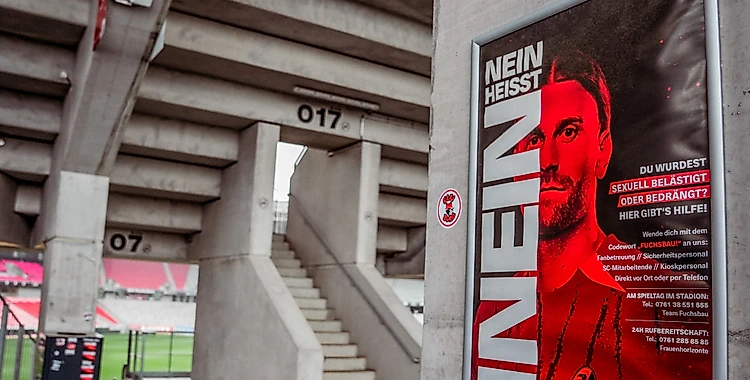 Plakat im Stadion mit der Aufschrift 'Nein heißt Nein'. Das Plakat zeigt ein Gesicht in Rot und Weiß und informiert über Hilfsangebote bei sexueller Belästigung oder Bedrängung. Im Hintergrund sind Treppen und Sitzplätze des Stadions zu sehen.