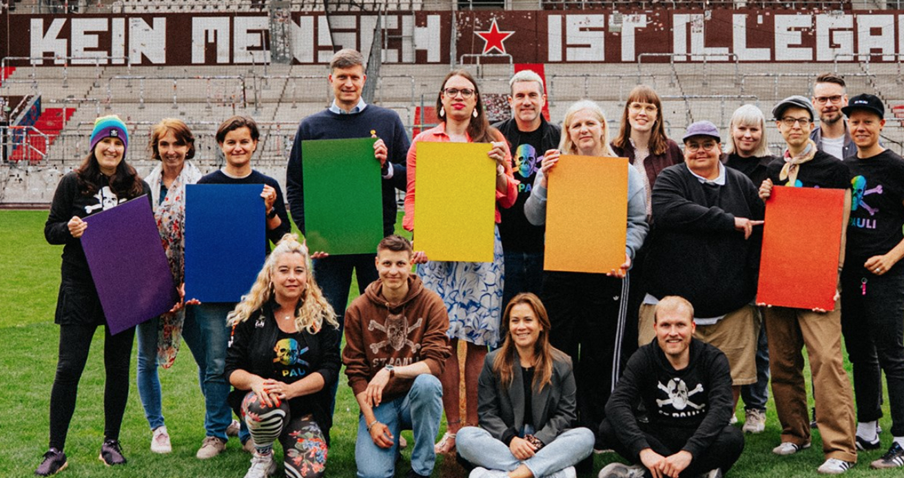Eine Gruppe von Menschen steht und kniet auf einem Fußballfeld. Sie halten bunte Pappkarten in den Farben lila, blau, grün, gelb, orange und rot. Im Hintergrund ist eine Wand mit der Aufschrift "KEIN MENSCH IST ILLEGAL" und einem roten Stern zu sehen. Die Gruppe scheint sich für ein gemeinsames Foto zu versammeln, möglicherweise im Rahmen einer Aktion oder eines Events zur Unterstützung von sozialen oder politischen Themen.