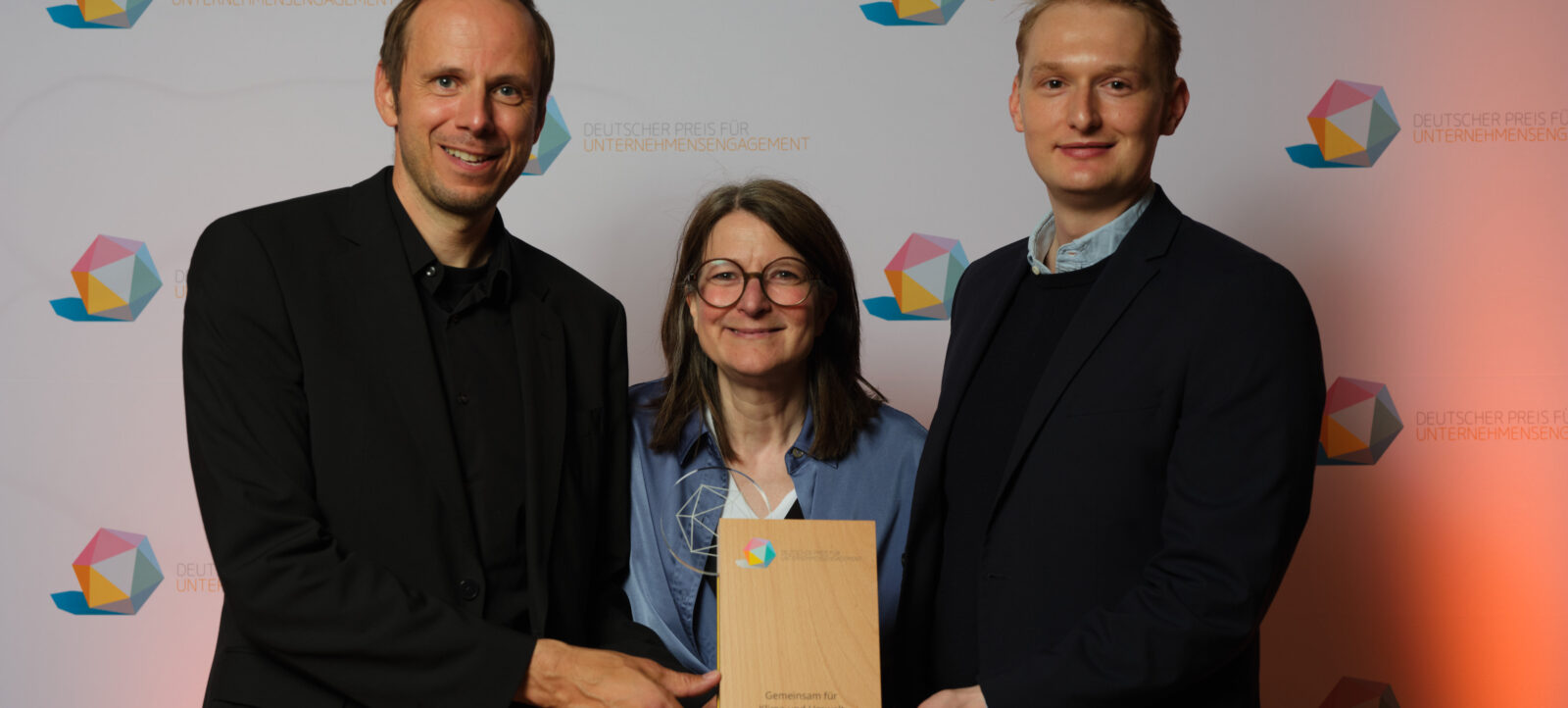 Drei Personen bei einer Preisverleihung des Deutschen Preises für Unternehmensengagement. Die mittlere Person hält einen Holzpreis in den Händen, während die beiden anderen Personen sie begleiten. Der Hintergrund zeigt das Logo des Deutschen Preises für Unternehmensengagement.