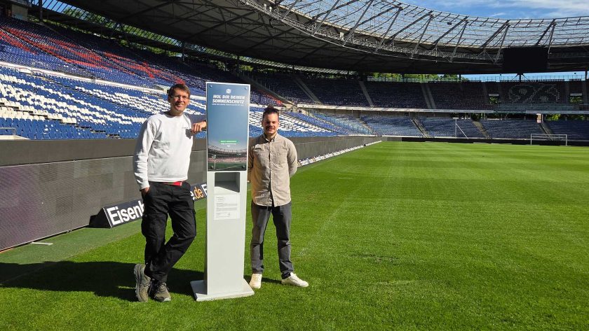 Zwei Männer stehen auf einem Fußballfeld und präsentieren einen Sonnencreme-Spender. Im Hintergrund sind die leeren Tribünen des Stadions zu sehen.