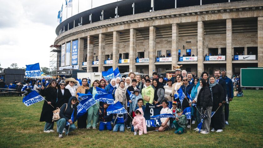Eine Gruppe von Menschen, darunter Frauen und Kinder, steht vor einem Fußballstadion und hält blaue Fahnen mit weißer Aufschrift. Sie lächeln und wirken fröhlich, während sie für ein Gruppenfoto posieren. Im Hintergrund ist das Stadion mit verschiedenen Werbebannern und Flaggen zu sehen. Die Szene findet auf einem Rasenbereich vor dem Stadion statt.