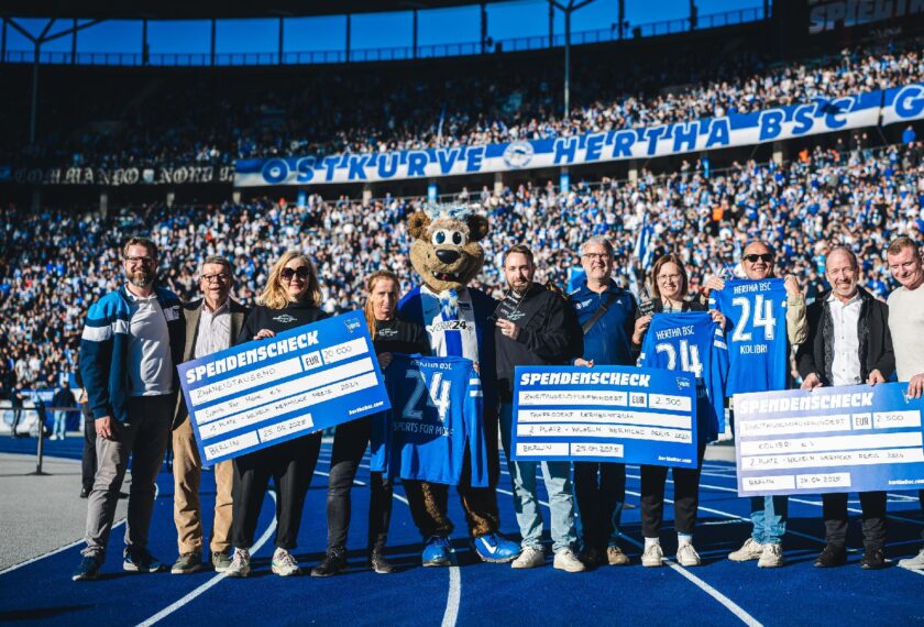 Gruppenfoto von Spendenübergabe auf dem Fußballfeld mit Hertha-BSC-Maskottchen und Fans im Hintergrund.