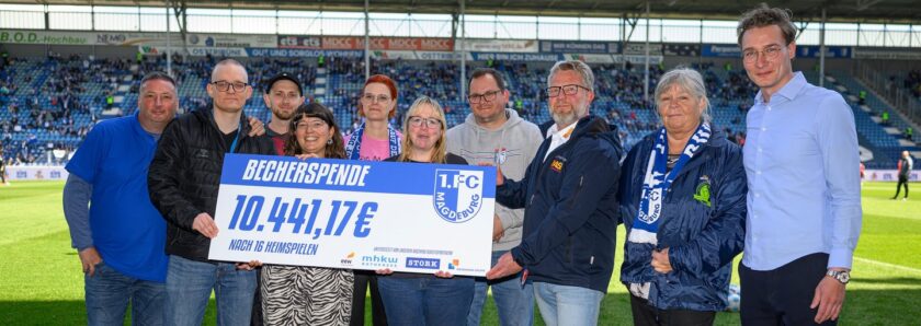 Eine Gruppe von Menschen steht auf einem Fußballfeld und hält ein großes Schild mit der Aufschrift "Becherspende 10.441,17€ nach 16 Heimspielen" und dem Logo des 1. FC Magdeburg. Die Personen lächeln und tragen verschiedene Kleidungsstücke, darunter Jacken und Schals in den Vereinsfarben. Im Hintergrund sind die Tribünen des Stadions zu sehen.