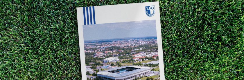 Ein Nachhaltigkeitsbericht des 1. FC Magdeburg liegt auf einem grünen Rasen. Auf dem Cover des Berichts ist eine Luftaufnahme des Stadions und der umliegenden Stadt zu sehen. Das Logo des 1. FC Magdeburg ist oben rechts auf dem Cover platziert.
