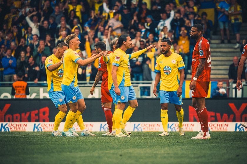 Fußballspieler im gelb-blauen Trikot feiern ein Tor, während ein Spieler im roten Trikot in der Nähe steht. Die Szene spielt auf dem Spielfeld während eines Spiels, mit begeisterten Fans im Hintergrund.