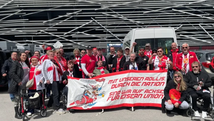 Eine Gruppe von Menschen, darunter einige im Rollstuhl, posieren vor einem Stadion. Sie halten ein Transparent mit der Aufschrift "Mit unseren Rollis durch die Nation sind wir auf Achse für Eisern Union". Die meisten tragen rote und weiße Kleidung, die mit dem Logo des Vereins Union Berlin verziert ist. Im Hintergrund sind ein Transporter und das Stadion zu sehen.
