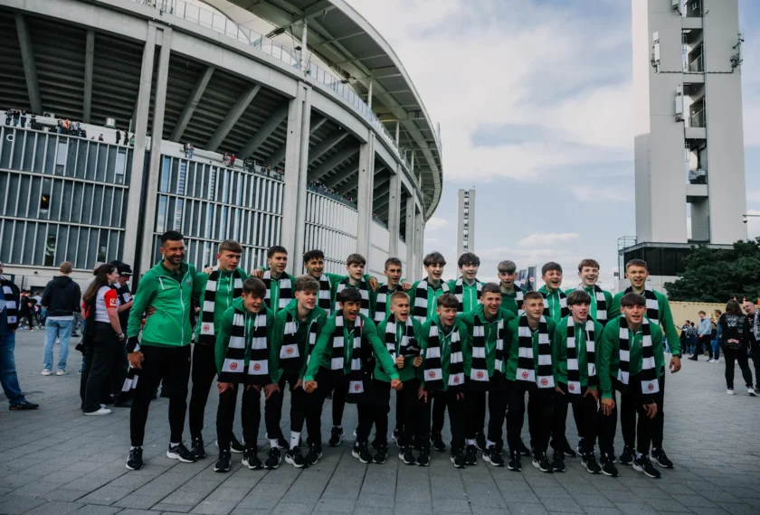 Eine Gruppe junger Fußballspieler in grün-schwarzen Trikots posiert vor einem großen Stadion. Die Spieler lächeln und scheinen sich für ein Gruppenfoto aufzustellen. Im Hintergrund sind weitere Personen und das moderne Stadiongebäude mit seiner architektonischen Struktur zu sehen.