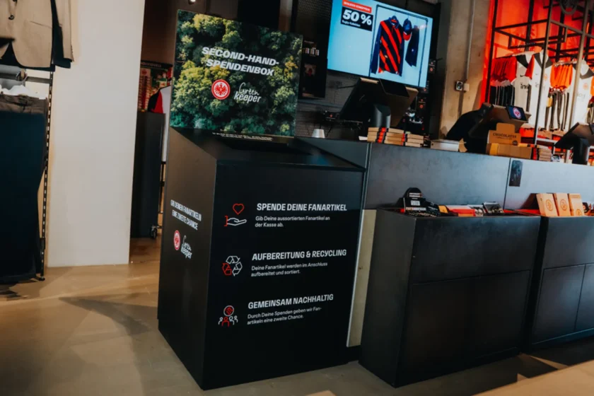 Second-Hand-Spendenbox im Eintracht Frankfurt Merchandising-Store. Die Box lädt Fans ein, ihre aussortierten Fanartikel abzugeben. Diese werden anschließend aufbereitet und recycelt, um gemeinsam nachhaltig zu handeln und den Fanartikeln eine zweite Chance zu geben. Im Hintergrund sind verschiedene Merchandising-Artikel und ein Bildschirm mit einem Angebot zu sehen.