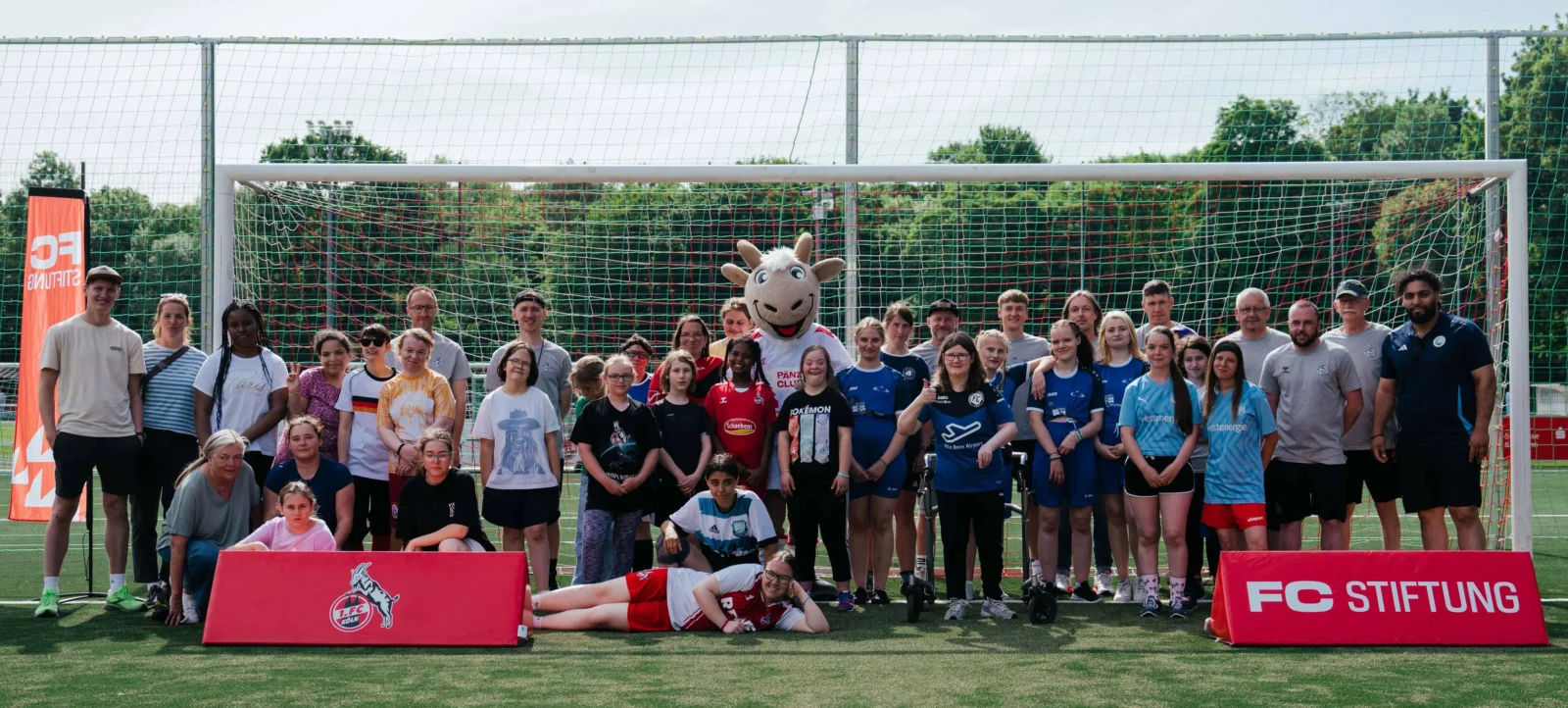 Eine Gruppe von Menschen, darunter Kinder und Erwachsene, steht vor einem Fußballtor auf einem Rasenplatz. Sie tragen verschiedene T-Shirts und Sportkleidung. In der Mitte der Gruppe steht eine Person im Kostüm eines Maskottchens. Vor dem Tor liegen zwei Personen auf dem Boden. Im Hintergrund ist ein Zaun und eine Werbebande mit dem Logo "FC STIFTUNG" zu sehen.