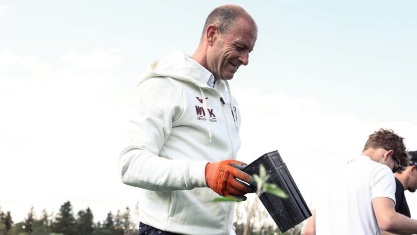 Ein Mann in einem weißen Hoodie mit dem Aufdruck "WIK Versicherungen" hält eine kleine Pflanze in einem schwarzen Topf. Er trägt orangefarbene Handschuhe und lächelt, während er die Pflanze betrachtet. Im Hintergrund sind weitere Personen und Bäume zu sehen.
