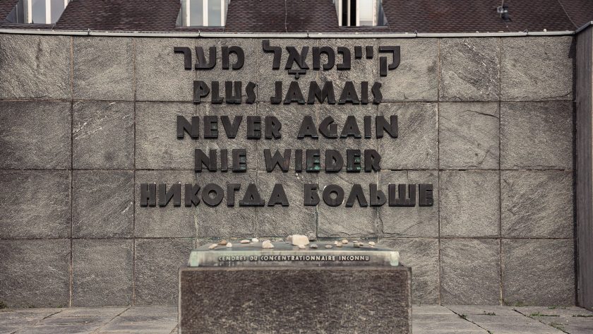 Ein Mahnmal mit der Inschrift "Plus jamais", "Never again", "Nie wieder" und "Никогда больше" in mehreren Sprachen, darunter Hebräisch, Französisch, Englisch, Deutsch und Russisch. Vor dem Mahnmal liegt eine Urne mit der Aufschrift "Cendres du concentrationnaire inconnu", was auf Französisch "Asche des unbekannten Häftlings" bedeutet. Das Mahnmal erinnert an die Opfer des Holocaust und die Bedeutung, solche Gräuel nie wieder zuzulassen.