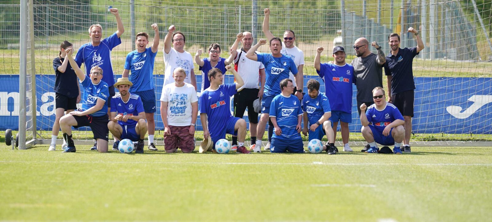 Eine Gruppe von Menschen in verschiedenen Trikots und Sportkleidung posiert vor einem Fußballtor. Die meisten von ihnen tragen blaue Trikots mit dem Logo von SAP und TSG Hoffenheim, während einige weiße T-Shirts mit Aufdrucken tragen. Sie sind in einer Mischung aus stehenden und knienden Positionen angeordnet und zeigen Freude und Begeisterung, einige mit erhobenen Fäusten und lachenden Gesichtern. Im Hintergrund ist ein Zaun mit Werbung zu sehen, und das Bild vermittelt einen Eindruck von Teamgeist und Inklusion während eines Fußballtrainings oder -events.