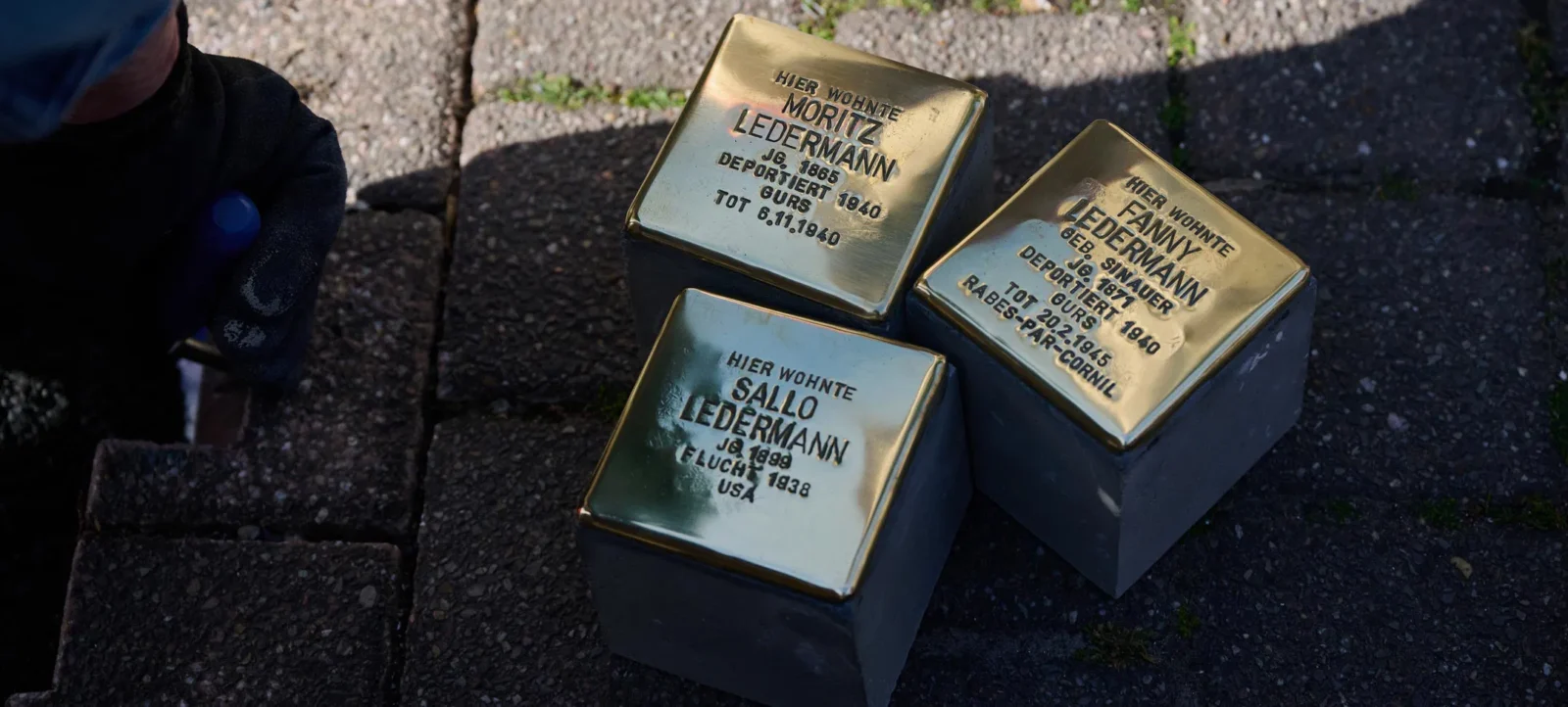 Drei goldene Stolpersteine auf einem Gehweg. Jeder Stein trägt die Inschrift mit dem Namen und den Lebensdaten einer Person, die hier gewohnt hat. Die Steine sind in den Boden eingelassen und erinnern an die Opfer des Nationalsozialismus. Eine Person in schwarzer Kleidung und Handschuhen ist dabei, einen der Steine zu reinigen.