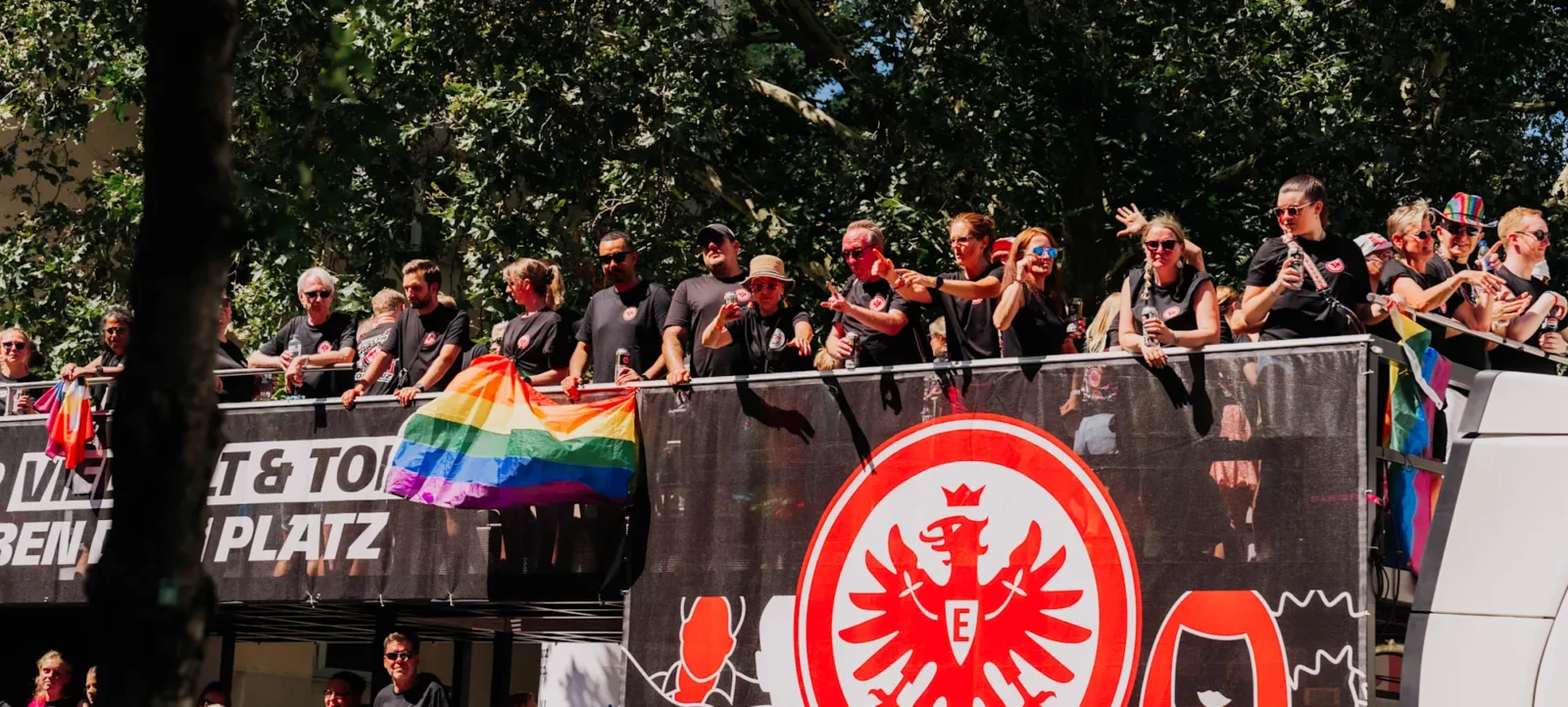 Eine Gruppe von Menschen steht auf einem Doppelstockbus und hält eine Regenbogenfahne. Sie tragen schwarze T-Shirts mit einem roten Adler-Logo und sind Teil einer Parade. Im Hintergrund sind Bäume und weitere Menschen zu sehen. Der Bus ist mit dem Schriftzug "VIER VIER TOR & TOR PLATZ" und dem Adler-Logo verziert.