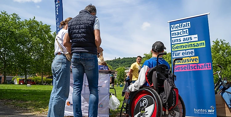 Gruppe von Menschen bei einem inklusiven Sporttag. Eine Person im Rollstuhl steht neben einem Banner, auf dem 'Vielfalt macht uns aus – Gemeinsam für eine inklusive Gesellschaft!' steht. Die Veranstaltung findet im Freien statt, mit Bäumen und Zelt im Hintergrund.