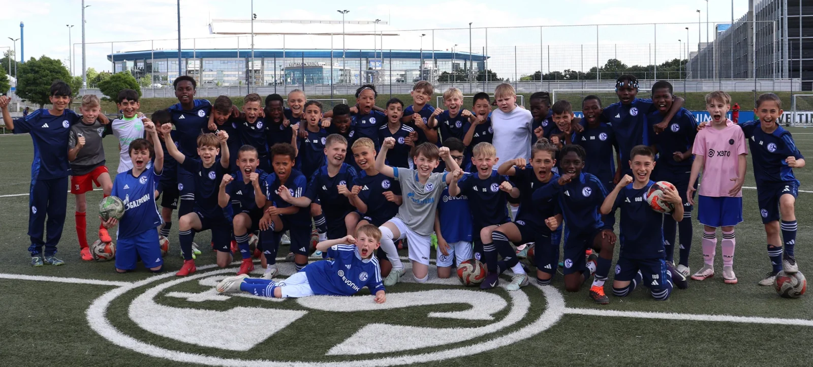 Eine Gruppe von Kindern und Jugendlichen in blauen Fußballtrikots posiert auf einem Fußballfeld. Sie stehen und knien um ein großes, weißes "9" auf dem Rasen. Die Kinder wirken fröhlich und energiegeladen, einige halten Fußballschuhe oder Bälle. Im Hintergrund ist ein modernes Stadion zu sehen.